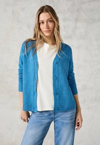 Ljusblå cardigan i mjuk ull med rund halsringning, långa ärmar och knäppning med knappar. Bärs över en vit topp med jeans i denim.