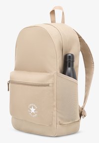 Beež kangast seljakott ümarate ülaosadega, ees lukuga tasku, võrkvõrguga küljetasku, kus must joogipudel sees, ja valge Converse logo detail.