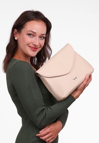 Beige kožená kabelka s uzávěrem na klopu, s lesklým stříbrným logem, kterou drží osoba v zeleném žebrovaném topu na bílém pozadí.