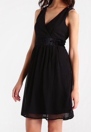 Donna che indossa un vestito nero senza maniche con scollo a V e fascia in velluto, in piedi con le braccia rilassate lungo i fianchi su uno sfondo uniforme.