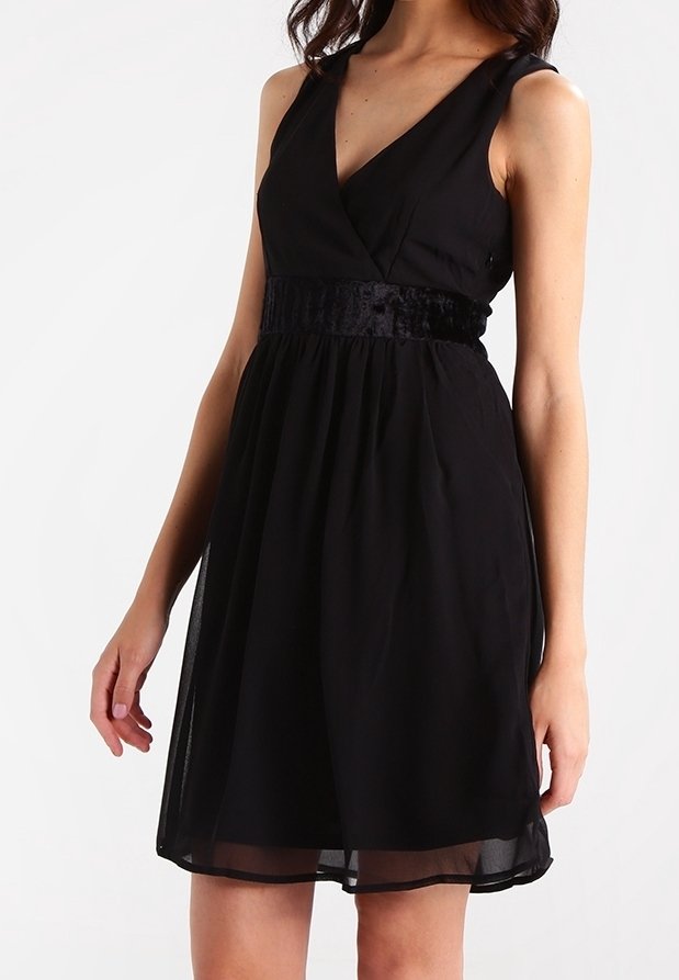 Femme portant une robe noire sans manches avec un col en V et une ceinture en velours, debout les bras détendus le long du corps, sur un fond uni.