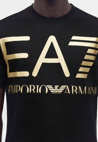 Μαύρο μπλουζάκι με μεγάλα χρυσά γράμματα "EA7" και λογότυπο "EMPORIO ARMANI" στο στήθος, σε άτομο με σκουρόχρωμο δέρμα.