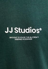 Σκούρο πράσινο ύφασμα με λευκό κείμενο "JJ Studios" και μικρότερο κείμενο "Brought to you by the J&J Crew, designed in Denmark" τυπωμένα πάνω του.