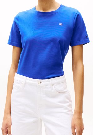 Osoba nosząca gładki królewsko niebieski t-shirt z krótkim rękawem, schowany w białe jeansy z wysokim stanem na białym tle.