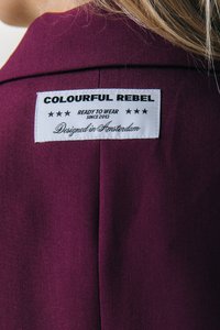 Maroon fabric jacket with a white label on the back. Label reads "COLOURFUL REBEL," features text "READY TO WEAR" and "Designed in Amsterdam."