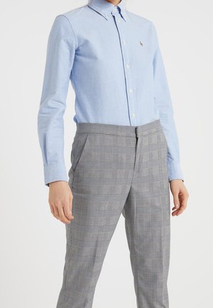 Personne portant une chemise bleue claire boutonnée avec un petit logo et un pantalon taille haute gris à carreaux, debout devant un fond blanc.
