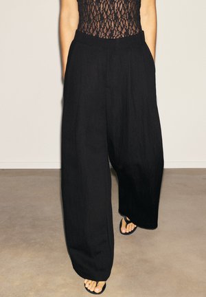 Personne portant un pantalon noir à jambes larges, un top en dentelle noir et des sandales noires type tongs, debout contre un mur uni.