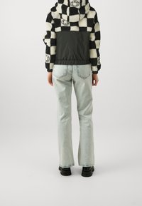 Persona vistiendo jeans rectos de color azul claro y una chaqueta a cuadros en blanco y negro con caras sonrientes, de pie sobre un fondo liso.