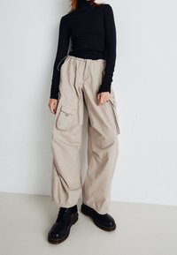 Beige Cargo-Hosen mit lockerer Passform und elastischem Bund, ausgestattet mit Seitentaschen und Kordelzugdetails. Schwarze Schnürstiefel vervollständigen das Outfit.