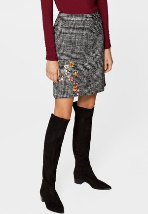 Kobieta w szarej mini spódnicy w kratę z kwiatowym haftem, bordowej bluzce z długim rękawem oraz czarnych zamszowych kozakach do kolan.