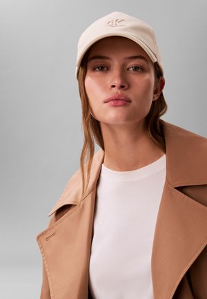 Beige pet met geborduurd logo, gedragen met een wit overhemd en een beige trenchcoat. Soepele stof met een ronde rand en subtiele stikdetails.