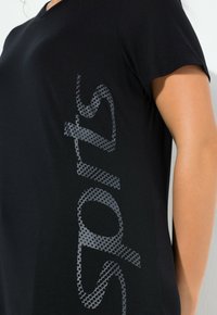 Camiseta negra de manga corta con un gran gráfico gris punteado de "sports" a lo largo del lateral. Tejido suave y liso con un corte holgado.