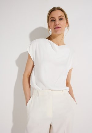 Femme aux cheveux blonds attachés, portant un chemisier blanc à manches courtes et un pantalon blanc taille haute, debout contre un mur blanc.