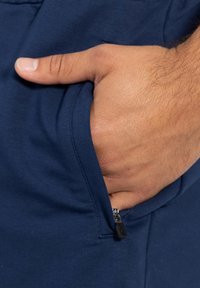 Prenda de color azul marino con un bolsillo con cremallera accesible; mano reposando dentro. Textura de tela suave, con un enfoque en primer plano en los detalles.