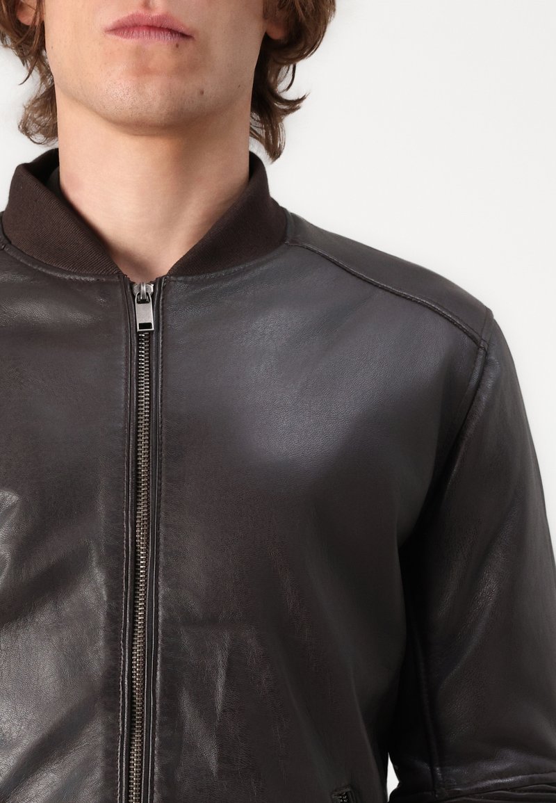 Donkerbruine leren bomberjack met een ritssluiting aan de voorkant, een geribbelde kraag en een gladde textuur. Beschikt over een getailleerd ontwerp en gestikte details.