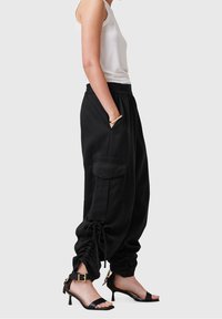 Femme portant un haut blanc sans manches, un pantalon cargo noir ample avec des détails à cordon de serrage, et des sandales à talons noires avec des boucles dorées.