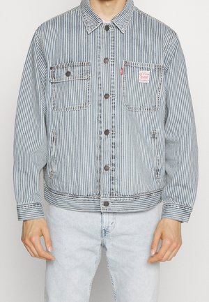 Mężczyzna ubrany w niebieską i białą, pionowo paskowaną dżinsową kurtkę z kieszeniami na piersi zapinanymi na guziki oraz jasnoniebieskie dżinsy, stoi z rozluźnionymi ramionami.