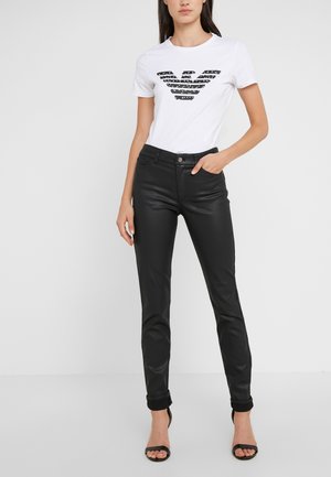 Femme portant un t-shirt blanc à manches courtes avec un motif noir et un pantalon en cuir noir ajusté, debout avec une main dans la poche et des talons noirs.