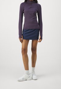 Fialový top s dlouhým rukávem a zipem u krku, spárovaný s modrou sukní. Nosený s bílými sportovními botami a ponožkami do kotníků. Hladký povrch po celé ploše.