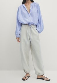 Chemise en lin bleu clair à manches longues avec une patte de boutonnage, associée à un pantalon ample gris clair avec une ceinture élastique. Tongs noires.