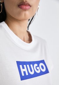 Gros plan d'une personne portant une chemise blanche avec un rectangle bleu et le mot "HUGO" imprimé sur la poitrine.