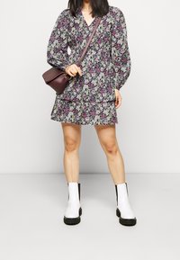 Kvinna klädd i en långärmad blommig klänning med vita ankelstövlar och en vinröd crossbody-väska, stående mot en enkel bakgrund.