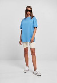 Femme portant un T-shirt bleu surdimensionné, un short de vélo crème, des baskets à plateforme blanches et des lunettes de soleil noires, debout devant un fond gris uni.