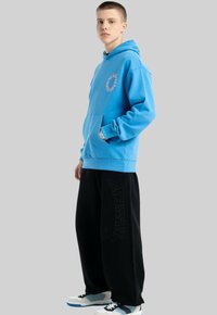 Jasnoniebieska bluza z kapturem z kieszenią kangurzą, ribbowymi mankietami i okrągłą grafiką na piersi. W zestawie z czarnymi spodniami z szerokimi nogawkami z logo.