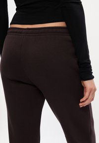 Brūnas sporta bikses ar elastīgu jostu, gluda tekstūra un sānu kabatām, kopā ar melnu garām piedurknēm kreklu.