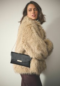 Cappotto in pelliccia sintetica color marrone chiaro, caratterizzato da una texture morbida. Viene indetta una pochette nera con motivo a coccodrillo, impreziosita da un accessorio in metallo dorato.