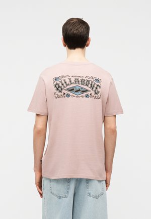 Άνδρας που φοράει ανοιχτό ροζ μπλούζα Billabong με μπλε και μαύρο λογότυπο στην πλάτη, σε συνδυασμό με ανοιχτό γαλάζιο τζιν σορτς, όρθιος με την πλάτη γυρισμένη.