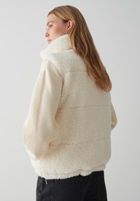 Crème-kleurige fleece gilet met hoge kraag, pluizige textuur en geribbelde crème mouwen. Heeft een ontspannen pasvorm en een lichte pof in het mouwontwerp.