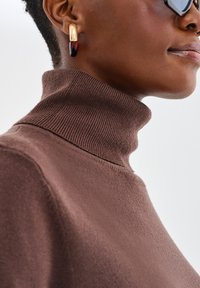 Brązowy sweter z golfem, wykonany z miękkiego materiału, z prążkowanym kołnierzem i bezszwowych ramionach, w połączeniu z złotą i burgundową kolczykiem.