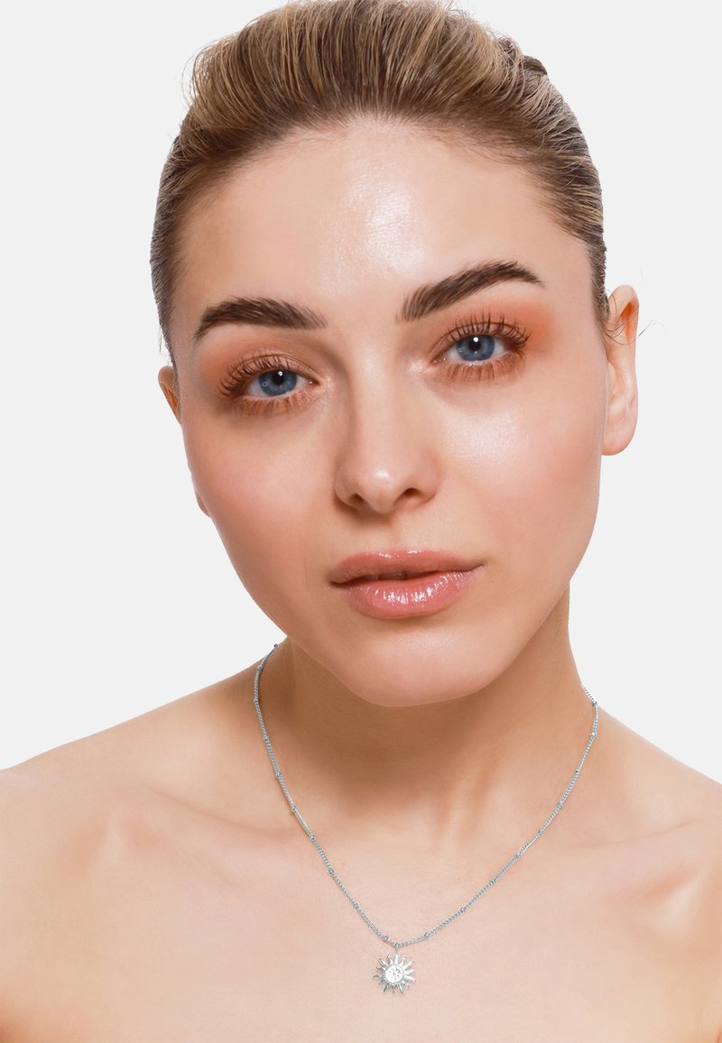 Srebrny naszyjnik z wisiorkiem w kształcie słońca, z wyraźnie zaznaczonymi promieniami i teksturowaną powierzchnią, noszony na nagiej szyi.