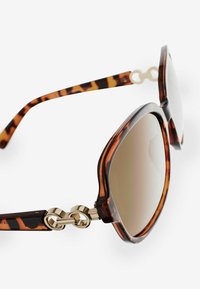 Gafas de sol de carey marrón con un diseño redondeado, detalles en dorado y lentes degradados. Presenta un detalle único de eslabones en las patillas.