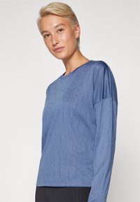 Maglietta blu a maniche lunghe con tessuto strutturato, caratterizzata da un colletto rotondo e maniche lisce a contrasto. Silhouette semplice e aderente.
