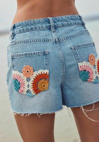 Denim lühikesed püksid, mille servad on katki, tagakotide peal on erksate heegeldatud lillemuster oranži, roosa ja teelrohelise värviga. Heleda sinise kangaga.