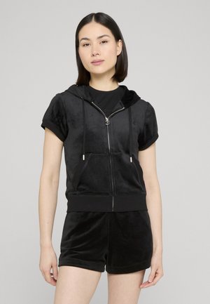 Kvinde iført sort kortærmet lynlås-hoodie med forlommer og matchende sorte shorts foran en ensfarvet grå baggrund.