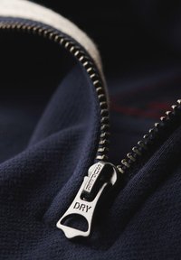 Tessuto blu navy con dettagli della cerniera in metallo argentato; il tirante della cerniera presenta l'incisione "SUPER DRY", denti esposti e texture liscia.