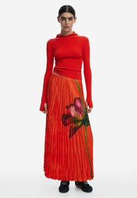 Pull à côtes rouges avec capuche, associé à une longue jupe plissée orange ornée d'un imprimé floral. Présenté sur un fond uni.
