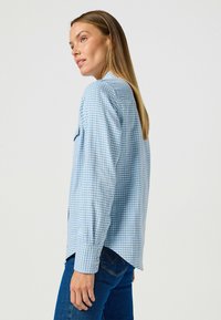 Niebieska koszula w kraciasty wzór z długimi rękawami, zapinanymi mankietami, spiczastym kołnierzykiem oraz luźnym fasonem, w połączeniu z ciemnoniebieskimi dżinsami.