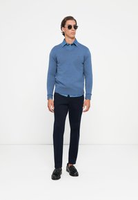Moški nosi sončna očala, modro V-izrezano pulover in jeans srajco, črne hlače in črne loafer čevlje, stoji pred belo ozadjem.