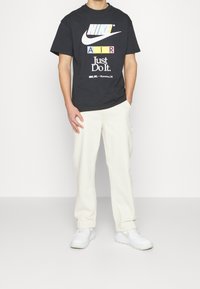 Personne portant un T-shirt graphique noir Nike Air, un pantalon blanc et des baskets blanches, se tenant avec une main dans la poche devant un fond uni.