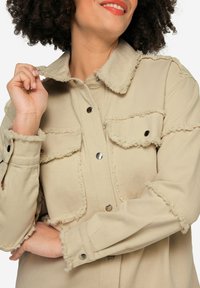 Giubbotto di denim beige con bordi sfrangiati, bottoni a pressione e due tasche sul petto. Presenta un colletto e maniche lunghe.