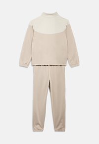 Σετ φόρμας σε μπεζ χρώμα που περιλαμβάνει πάνω με φερμουάρ, ψηλό γιακά και ελαστικές μανσέτες, συνδυασμένο με παντελόνι με ελαστική μέση στο ίδιο χρώμα. Λείο ύφασμα.
