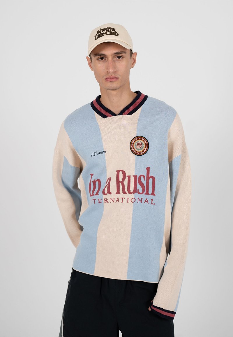 Homme portant un pull à rayures verticales beige et bleu clair avec le texte « In a Rush INTERNATIONAL » et une casquette beige avec le texte « Always Live Club ».