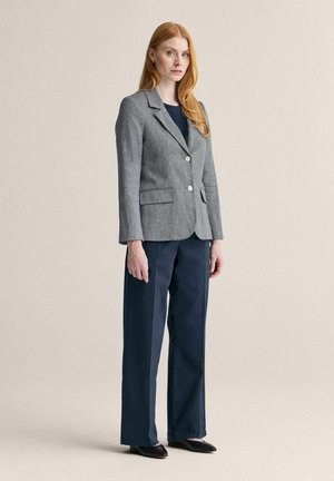 Kvinde med langt rødt hår iført en grå blazer, sort top, marineblå bukser med vide ben og sorte flade sko, stående mod en beige baggrund.