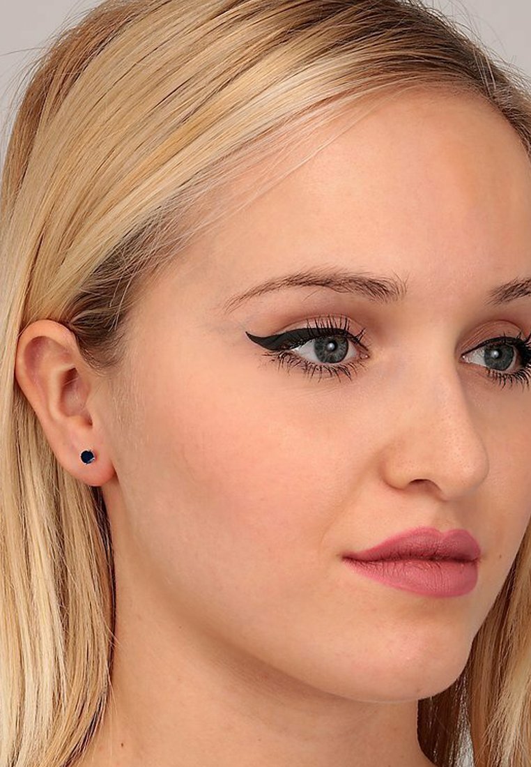 Blaue Ohrstecker in runder Form mit facettiertem Design. Das Modell hat glattes, blondes Haar und ein natürliches Make-up.