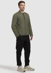 Olivenfarvet quilted jakke med opretstående krave, frontlynlås og sidelommer; parret med sorte cargo bukser og lyse sneakers.