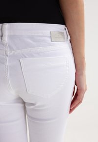 Jeans de denim blanco con una textura suave y bolsillos en la parte trasera. Presenta una etiqueta de marca en la cinturilla, mostrando el nombre de la marca.